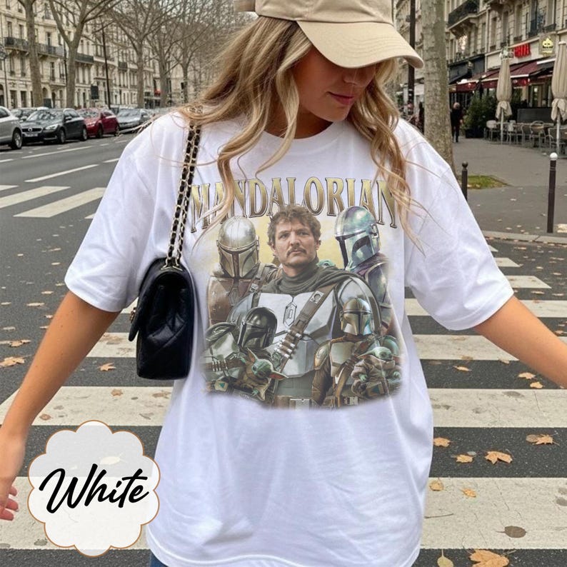 Pu&ograve; includere: Una t-shirt bianca a maniche corte con una stampa grafica di personaggi di The Mandalorian, tra cui una figura centrale e diverse figure in armatura. Il testo "MANDALORIAN" &egrave; stampato in un carattere color oro sopra l'illustrazione.