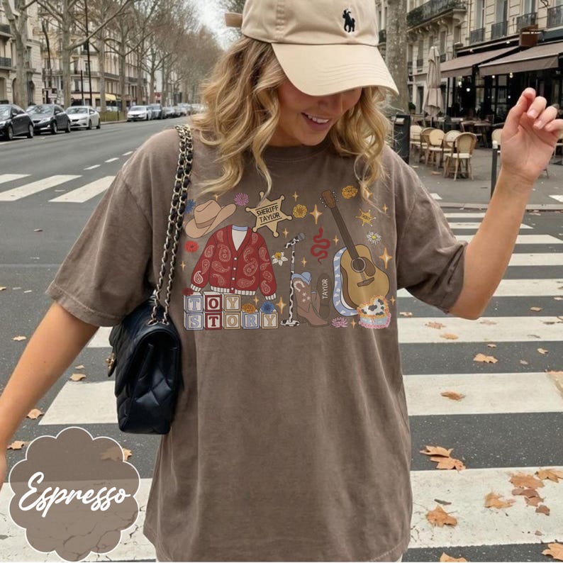 May include: A brown short-sleeve t-shirt featuring a graphic print with a cowboy hat, a red paisley cardigan, a sheriff badge labeled Sheriff Taylor, a microphone, a guitar, and blocks spelling Toy Story. A black quilted shoulder bag is worn.