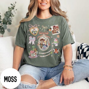 May include: Moss green t-shirt featuring a collection of cartoon character graphics. The design includes images of Woody, Buzz Lightyear, and other characters. The word "MOSS" is printed on a white circle at the bottom left.