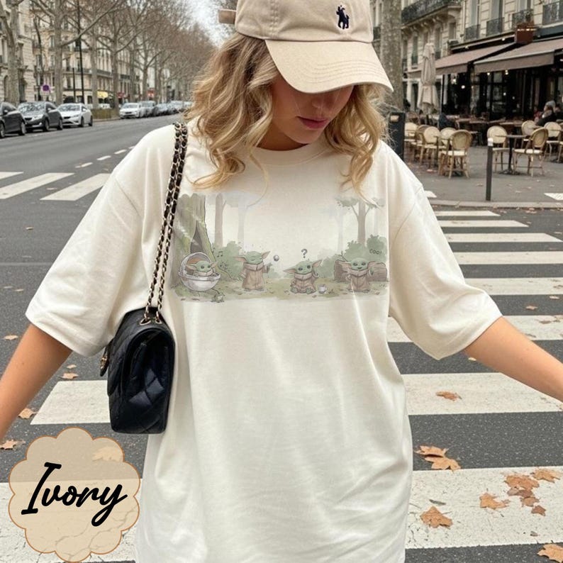 Puede incluir: Camiseta color marfil con un gr&aacute;fico de varios personajes de Grogu en un entorno forestal. La camiseta tiene mangas cortas y un corte holgado. La palabra "Ivory" est&aacute; escrita en cursiva en la parte inferior.