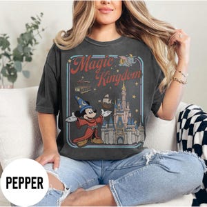 May include: Dark grey t-shirt with a vintage graphic of Mickey Mouse in a sorcerer's outfit, the Magic Kingdom logo, and a castle. The design includes a trolley, a ship, and stars. The shirt is worn by a person.