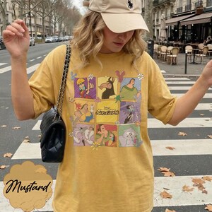 May include: A mustard yellow t-shirt featuring a grid of colorful illustrations from Disney's The Emperor's New Groove, including characters like Kuzco, Pacha, and Yzma, accented with small flower and mouse ear icons, paired with a black quilted shoulder bag.