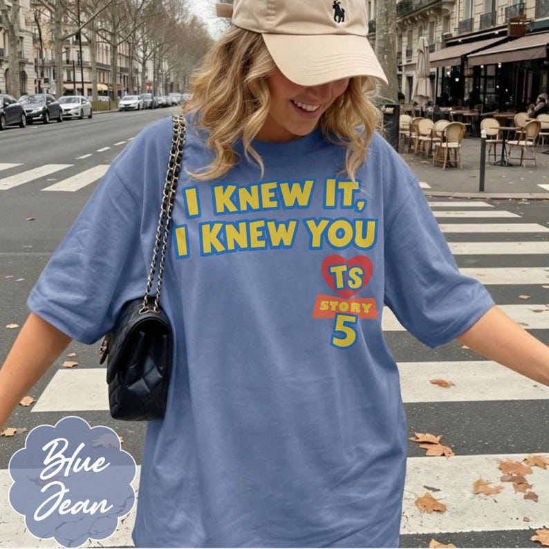 May include: A blue t-shirt featuring the yellow text I KNEW IT, I KNEW YOU, a red heart with the letters TS, and the word STORY above the number 5 in yellow. A black quilted handbag with a gold chain strap is worn over the shoulder.