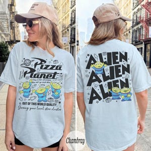 K&ouml;nnte beinhalten: Hellblaues T-Shirt mit Pizza Planet-Design und Alien-Charakteren. Auf der Vorderseite befindet sich das Pizza Planet-Logo und Text. Auf der R&uuml;ckseite steht das Wort "Alien" mehrfach. Ein l&auml;ssiges Kleidungsst&uuml;ck.