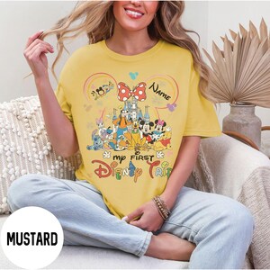 May include: Mustard-colored t-shirt featuring a colorful Disney graphic. The design includes Mickey and Minnie Mouse, a castle, and the text "my FIRST Disney TRIP". A red and white polka dot bow is at the top of the design. The word "Name" is also in the design.