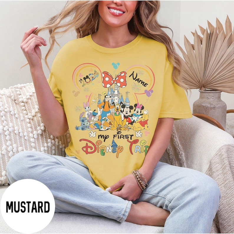 May include: Mustard-colored t-shirt featuring a colorful Disney graphic. The design includes Mickey and Minnie Mouse, a castle, and the text "my FIRST Disney TRIP". A red and white polka dot bow is at the top of the design. The word "Name" is also in the design.