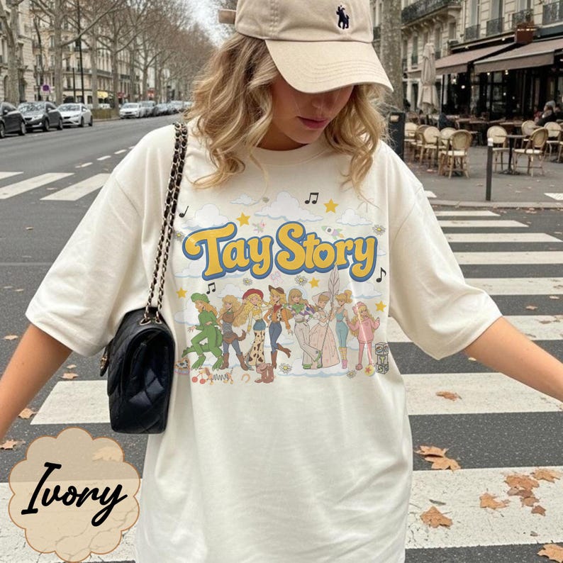 May include: An ivory short-sleeve t-shirt featuring a graphic print with the text Tay Story in yellow letters. The design includes a row of illustrated figures in various outfits, musical notes, stars, and clouds. A black quilted shoulder bag is present.