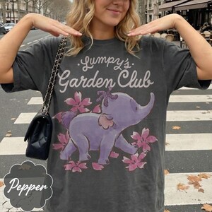 Op de afbeelding: Donkergrijze t-shirt met de tekst "Lumpy's Garden Club" boven een paarse olifantenillustratie omringd door roze bloemen. De t-shirt heeft een vintage uitstraling.