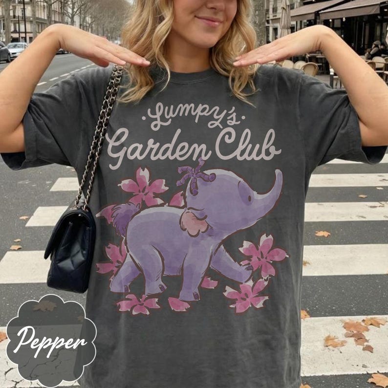 Op de afbeelding: Donkergrijze t-shirt met de tekst "Lumpy's Garden Club" boven een paarse olifantenillustratie omringd door roze bloemen. De t-shirt heeft een vintage uitstraling.