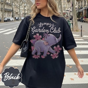 Op de afbeelding: Zwart T-shirt met de tekst "Lumpy's Garden Club" en een paarse olifantafbeelding omringd door roze bloemen. Het shirt heeft korte mouwen en een losse pasvorm. Het woord "Black" staat in witte letters op een zwarte achtergrond.