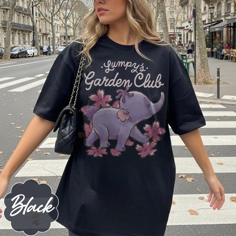 Op de afbeelding: Zwart T-shirt met de tekst "Lumpy's Garden Club" en een paarse olifantafbeelding omringd door roze bloemen. Het shirt heeft korte mouwen en een losse pasvorm. Het woord "Black" staat in witte letters op een zwarte achtergrond.