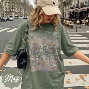 May include: A moss green t-shirt with a colourful graphic of Universal Studios attractions and characters. The shirt has short sleeves and a crew neck. The text "UNIVERSAL Studios" is printed on the front. A black purse with a chain strap is visible.