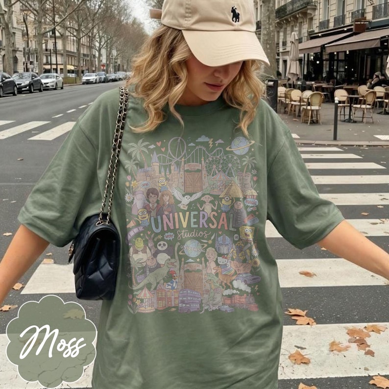 May include: A moss green t-shirt with a colourful graphic of Universal Studios attractions and characters. The shirt has short sleeves and a crew neck. The text "UNIVERSAL Studios" is printed on the front. A black purse with a chain strap is visible.