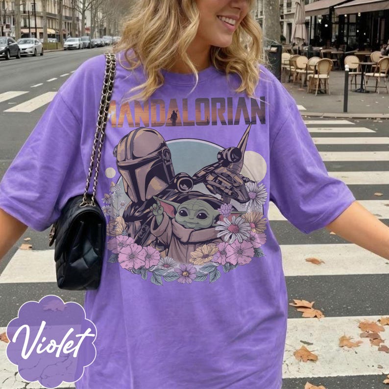 May include: Violet-colored t-shirt featuring a graphic of The Mandalorian, Grogu, and a spaceship, surrounded by flowers. The word "MANDALORIAN" is printed across the top. The shirt is a relaxed fit.