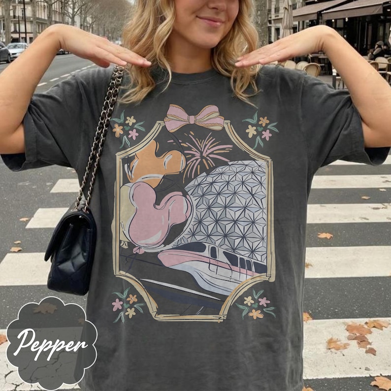 May include: Dark gray t-shirt with a colorful graphic featuring a pink bow, fireworks, a Mickey Mouse balloon, and a geometric sphere. The word "Pepper" is written in a decorative font at the bottom.