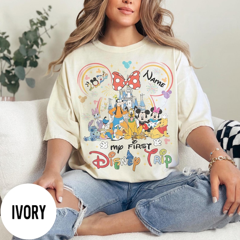 May include: Ivory-colored t-shirt featuring a colorful Disney graphic. The design includes Mickey and Minnie Mouse, Donald Duck, and other characters, with the text "my FIRST Disney TRIP" and a space for a name. The shirt is a relaxed fit.