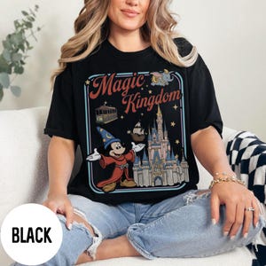 May include: Black t-shirt with a retro-style graphic of Mickey Mouse as a sorcerer, a castle, and the words "Magic Kingdom." The design includes a trolley, a ship, and Dumbo. The shirt is worn by a person.