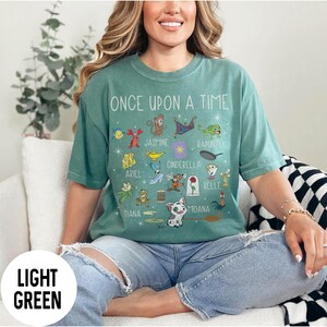 May include: Light green t-shirt with the text "ONCE UPON A TIME" and various cartoon character illustrations. The shirt features characters like Ariel, Jasmine, Cinderella, and Moana. The shirt is a light green colour.