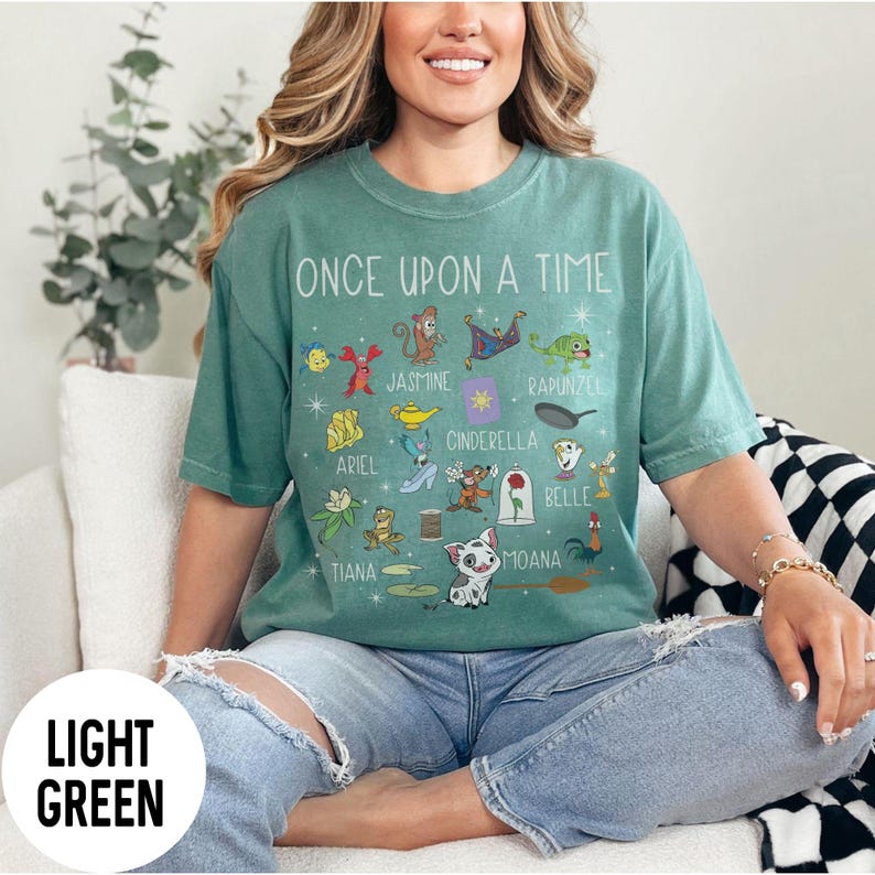 May include: Light green t-shirt with the text "ONCE UPON A TIME" and various cartoon character illustrations. The shirt features characters like Ariel, Jasmine, Cinderella, and Moana. The shirt is a light green colour.