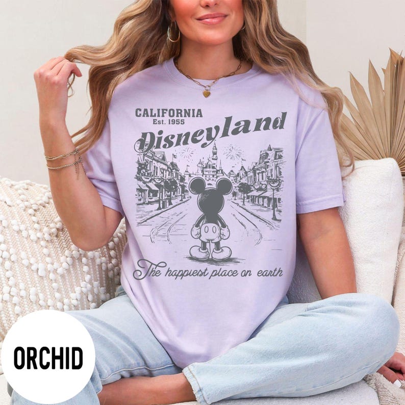 Może przedstawiać: Jasnofioletowa koszulka z grafiką Disneyland. Grafika przedstawia Myszka Miki przed sceną uliczną z napisem "California Est. 1955 Disneyland" i "The happiest place on earth."