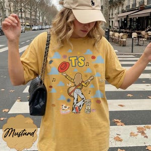 May include: A mustard yellow t-shirt featuring a graphic print of a person with long hair, a yellow plaid shirt, and cow-print pants, holding a guitar. The design includes a red hat, blue clouds, musical notes, flowers, and blocks with the letters TS.