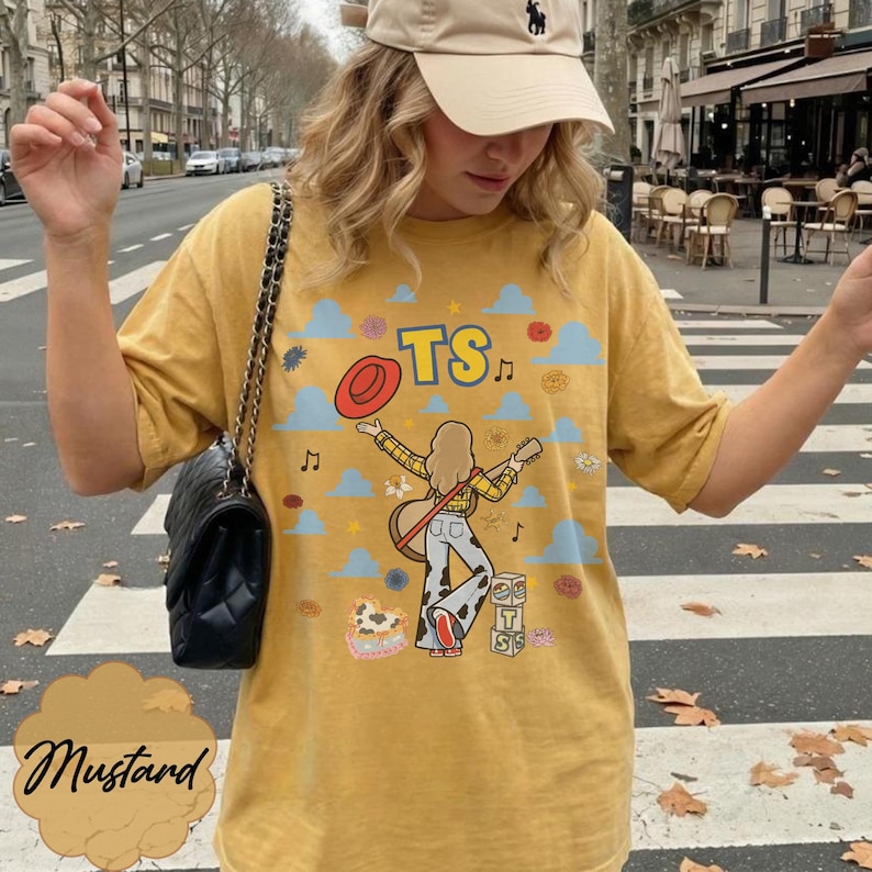 May include: A mustard yellow t-shirt featuring a graphic print of a person with long hair, a yellow plaid shirt, and cow-print pants, holding a guitar. The design includes a red hat, blue clouds, musical notes, flowers, and blocks with the letters TS.