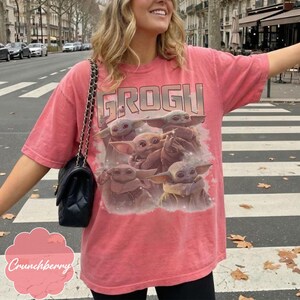 May include: A coral-colored short-sleeve t-shirt featuring a graphic print of multiple small green creatures with large ears and eyes. The text GROGU is printed in a bold, blocky font above the creatures. The shirt is paired with a black quilted shoulder bag.