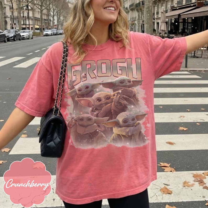 May include: A coral-colored short-sleeve t-shirt featuring a graphic print of multiple small green creatures with large ears and eyes. The text GROGU is printed in a bold, blocky font above the creatures. The shirt is paired with a black quilted shoulder bag.