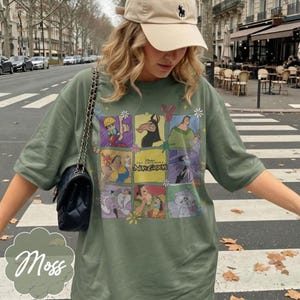 May include: An olive green t-shirt featuring a grid of colorful graphic illustrations from the movie The Emperor's New Groove, including characters like Kuzco, Pacha, and Yzma, with the movie title text centered in the middle of the design.
