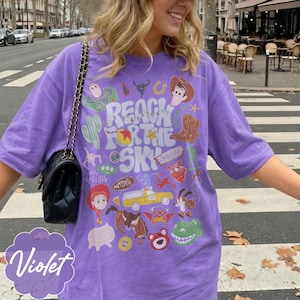 Pode incluir: Camiseta roxa com um design gr&aacute;fico com personagens e frases de um filme de anima&ccedil;&atilde;o popular. O design inclui um cacto, botas de cowboy e a frase "Reach for the Sky."