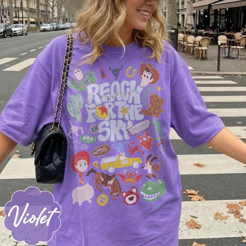 Pode incluir: Camiseta roxa com um design gr&aacute;fico com personagens e frases de um filme de anima&ccedil;&atilde;o popular. O design inclui um cacto, botas de cowboy e a frase "Reach for the Sky."