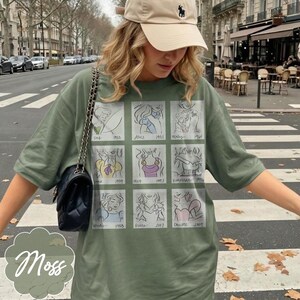 May include: Moss green t-shirt featuring a grid of cartoon illustrations with dates. The shirt has a crew neck and short sleeves. A black purse with a chain strap hangs on the left side. A beige baseball cap is worn.