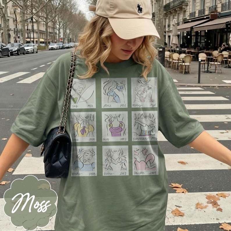 May include: Moss green t-shirt featuring a grid of cartoon illustrations with dates. The shirt has a crew neck and short sleeves. A black purse with a chain strap hangs on the left side. A beige baseball cap is worn.