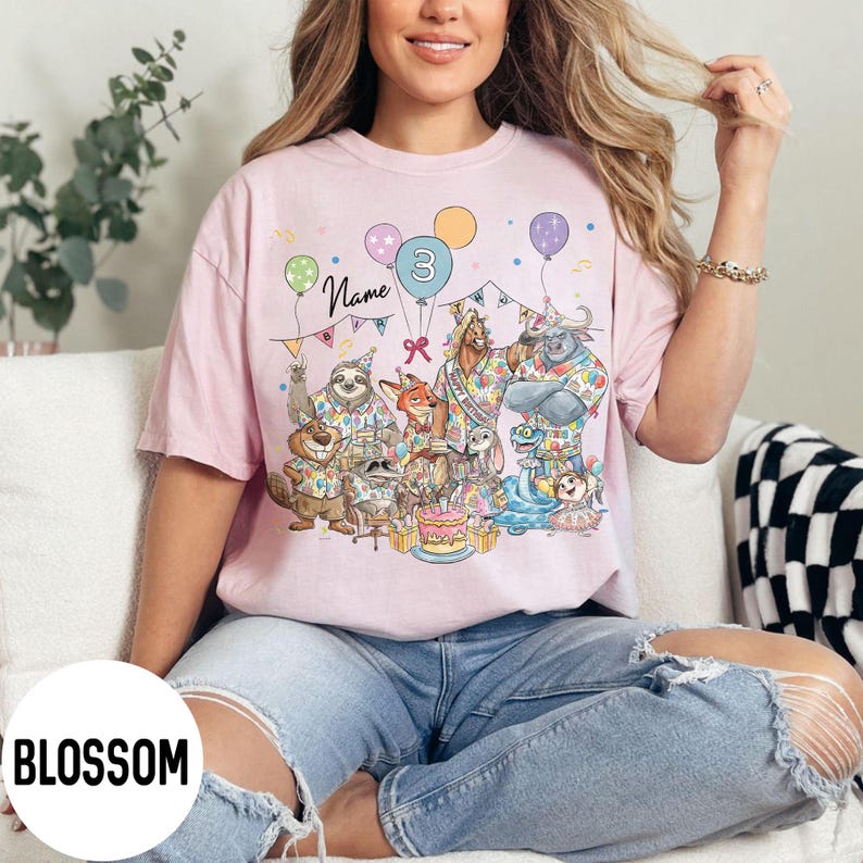 以下が含まれることがあります： 漫画の動物、風船、ケーキが描かれた、カラフルな誕生日パーティーのシーンが特徴の薄いピンク色のTシャツ。「3」の数字が風船に表示され、「Name」の文字が動物の上に書かれています。「BLOSSOM」の文字が白い円に印刷されています。