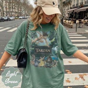 Op de afbeelding: Lichtgroen T-shirt met een Tarzan-afbeelding. Het ontwerp bevat het woord "Tarzan" in een rechthoekig kader, met illustraties van Tarzan en andere personages. Het T-shirt is lichtgroen van kleur.
