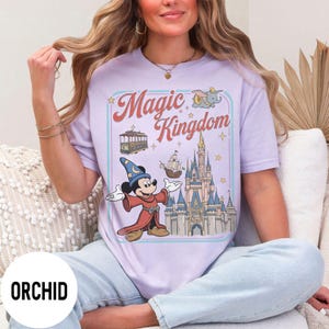 May include: Lilac t-shirt with a graphic of Mickey Mouse as a sorcerer in front of a castle, with the words "Magic Kingdom" in a retro design. The shirt also includes images of a cable car, a ship, and Dumbo the elephant.