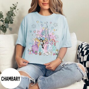 May include: Light blue t-shirt featuring a colorful graphic of a castle and several princesses. The design includes fireworks, balloons, and floral accents. The word "CHAMBARY" is printed in white on the bottom left of the shirt.