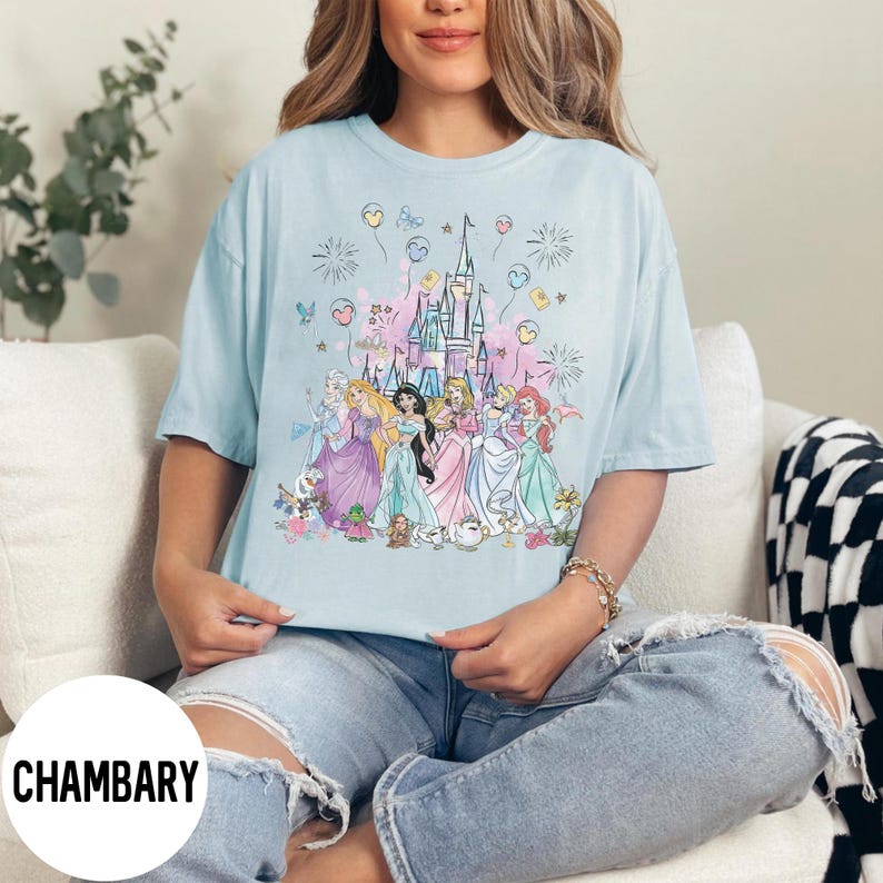 May include: Light blue t-shirt featuring a colorful graphic of a castle and several princesses. The design includes fireworks, balloons, and floral accents. The word "CHAMBARY" is printed in white on the bottom left of the shirt.