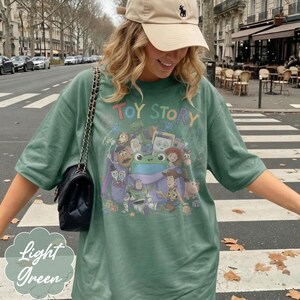 May include: A light green t-shirt featuring a graphic print of Toy Story characters including Woody, Buzz Lightyear, and a central green frog. The shirt has short sleeves and a crew neckline. A black quilted shoulder bag with a chain strap is worn.