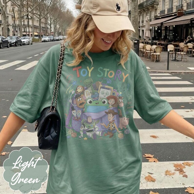 May include: A light green t-shirt featuring a graphic print of Toy Story characters including Woody, Buzz Lightyear, and a central green frog. The shirt has short sleeves and a crew neckline. A black quilted shoulder bag with a chain strap is worn.