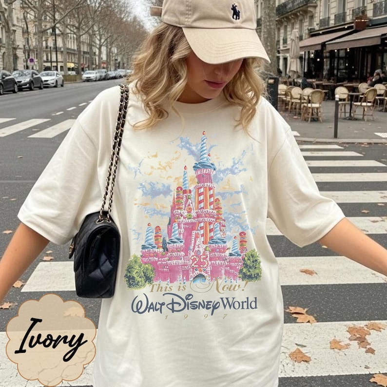 K&ouml;nnte beinhalten: Ein elfenbeinfarbenes Kurzarm-T-Shirt mit einer Grafik eines rosafarbenen Schlosses mit blauen Akzenten, dem Text "This is Now! Walt Disney World 1997" und einem "25"-Emblem. Eine schwarze gesteppte Umh&auml;ngetasche mit Goldkette wird &uuml;ber der Schulter getragen.