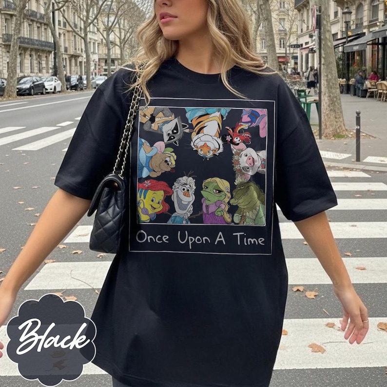 May include: Black t-shirt featuring a colorful graphic of cartoon characters with the text "Once Upon A Time." The shirt has short sleeves and a relaxed fit. The word "Black" is written in a decorative font on the bottom left.