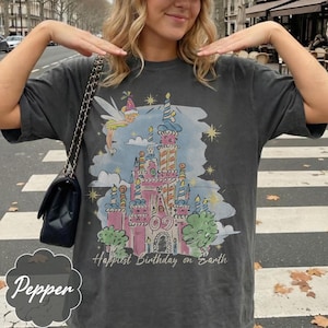 Puede incluir: Camiseta gris oscuro con una ilustraci&oacute;n de castillo colorido, un hada y el texto "Happiest Birthday on Earth". El dise&ntilde;o incluye elementos rosas, azules, amarillos y verdes. Es una prenda casual.