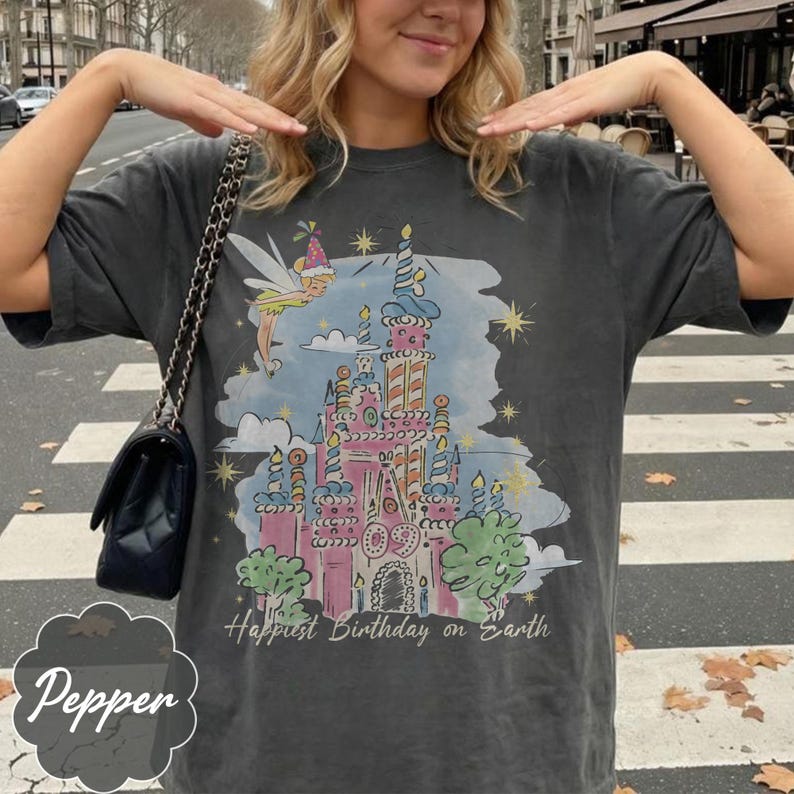 Puede incluir: Camiseta gris oscuro con una ilustraci&oacute;n de castillo colorido, un hada y el texto "Happiest Birthday on Earth". El dise&ntilde;o incluye elementos rosas, azules, amarillos y verdes. Es una prenda casual.