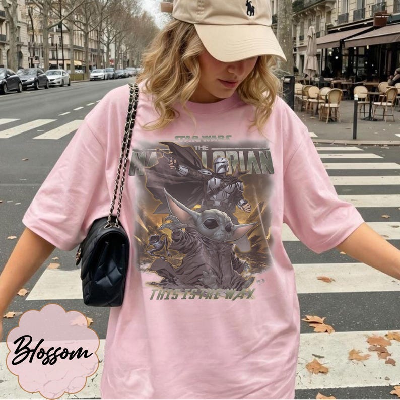 Puede incluir: Una camiseta rosa claro con un estampado gr&aacute;fico de The Mandalorian y Grogu de Star Wars. El dise&ntilde;o incluye el texto Star Wars, The Mandalorian y This Is The Way. Un bolso de hombro negro acolchado con correa de cadena se lleva sobre el hombro.