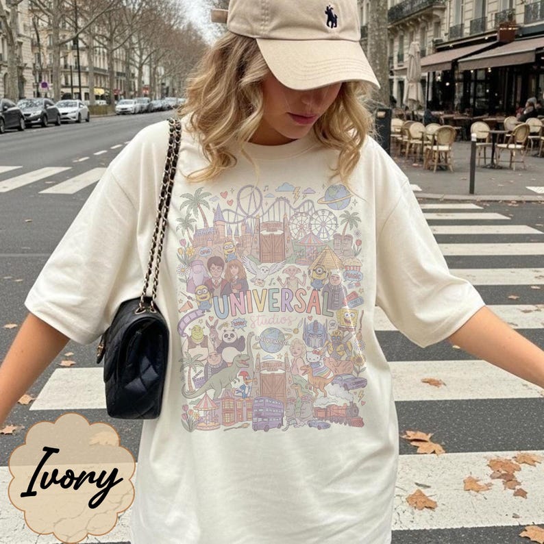 May include: Ivory t-shirt featuring a colourful graphic of Universal Studios characters and attractions. The shirt has the text "UNIVERSAL Studios". The person is wearing a beige cap and carrying a black purse with a chain strap.