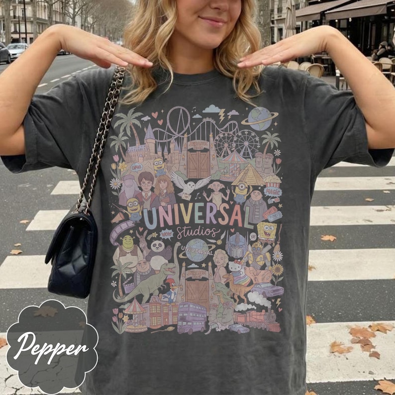 May include: Dark grey t-shirt with a vibrant graphic of Universal Studios characters and attractions. The design includes roller coasters, palm trees, and the text "UNIVERSAL Studios". A black handbag with a chain strap is visible.