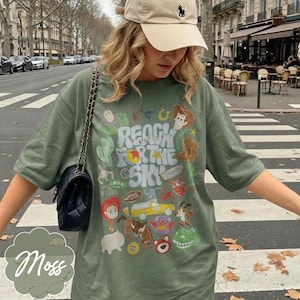 Pode incluir: T-shirt verde musgo com um design gr&aacute;fico colorido com personagens de desenhos animados e o texto "REACH FOR THE SKY". A camisa &eacute; combinada com um bon&eacute; de beisebol bege e uma bolsa preta.