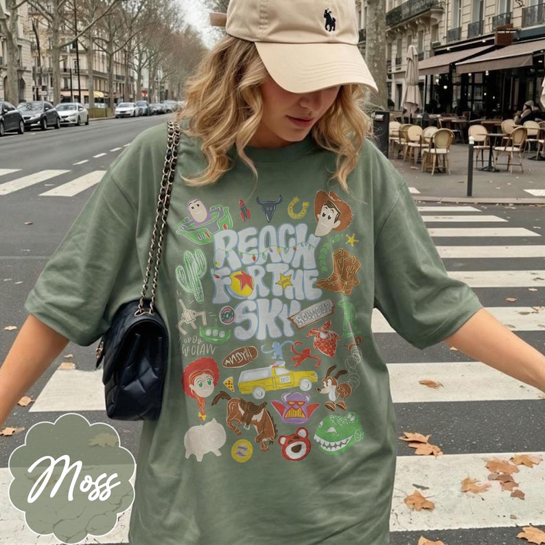 Pode incluir: T-shirt verde musgo com um design gr&aacute;fico colorido com personagens de desenhos animados e o texto "REACH FOR THE SKY". A camisa &eacute; combinada com um bon&eacute; de beisebol bege e uma bolsa preta.