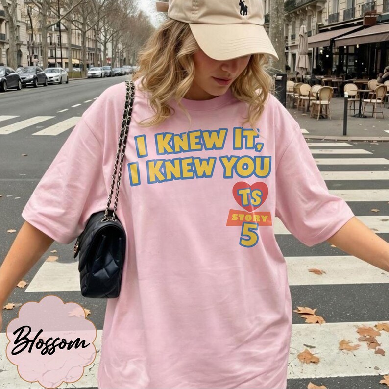 May include: A light pink short-sleeved t-shirt featuring the blue and yellow text I KNEW IT, I KNEW YOU, a red heart with the letters TS, and the word STORY above the number 5 in blue and yellow, paired with a black quilted shoulder bag.